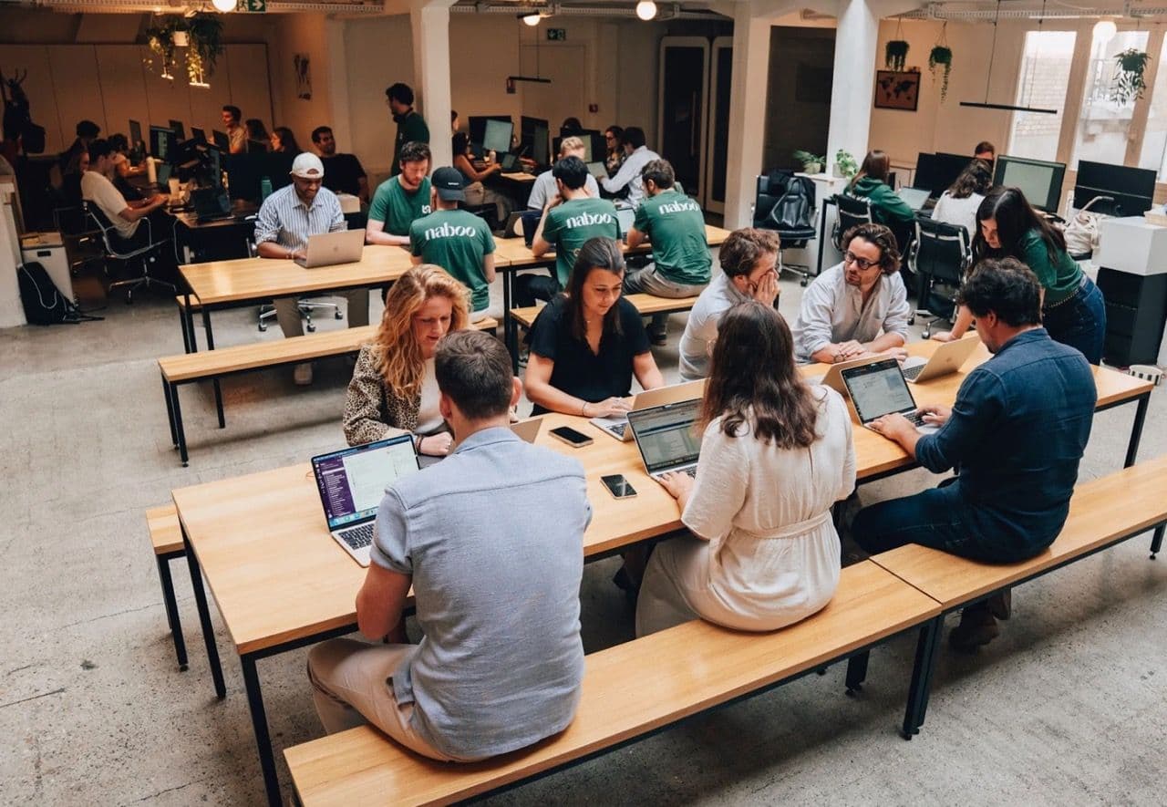 A large group works collaboratively on laptops at long wooden tables in a modern open-plan event space.