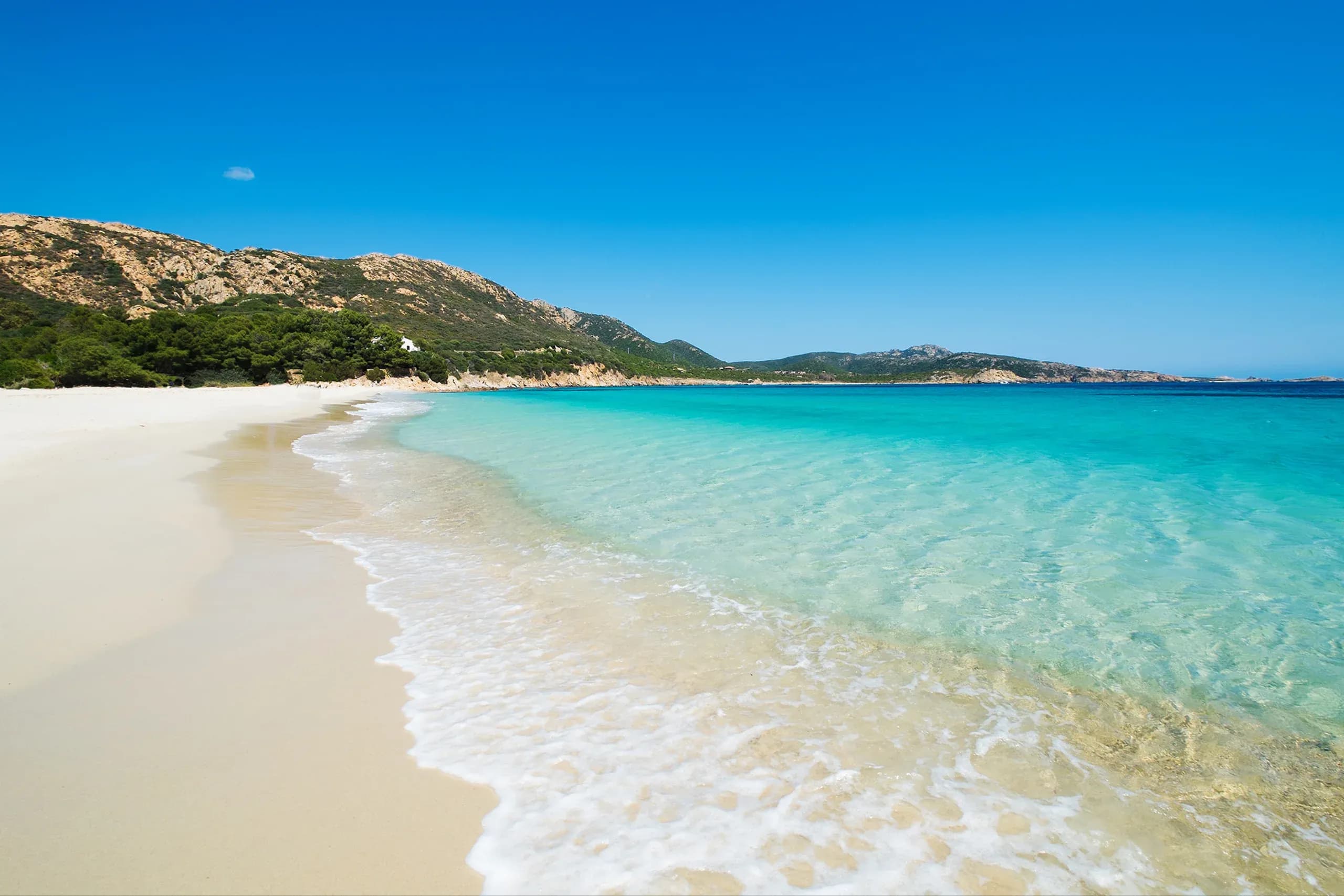 White sand beach with clear turquoise water and green hills under a bright blue sky