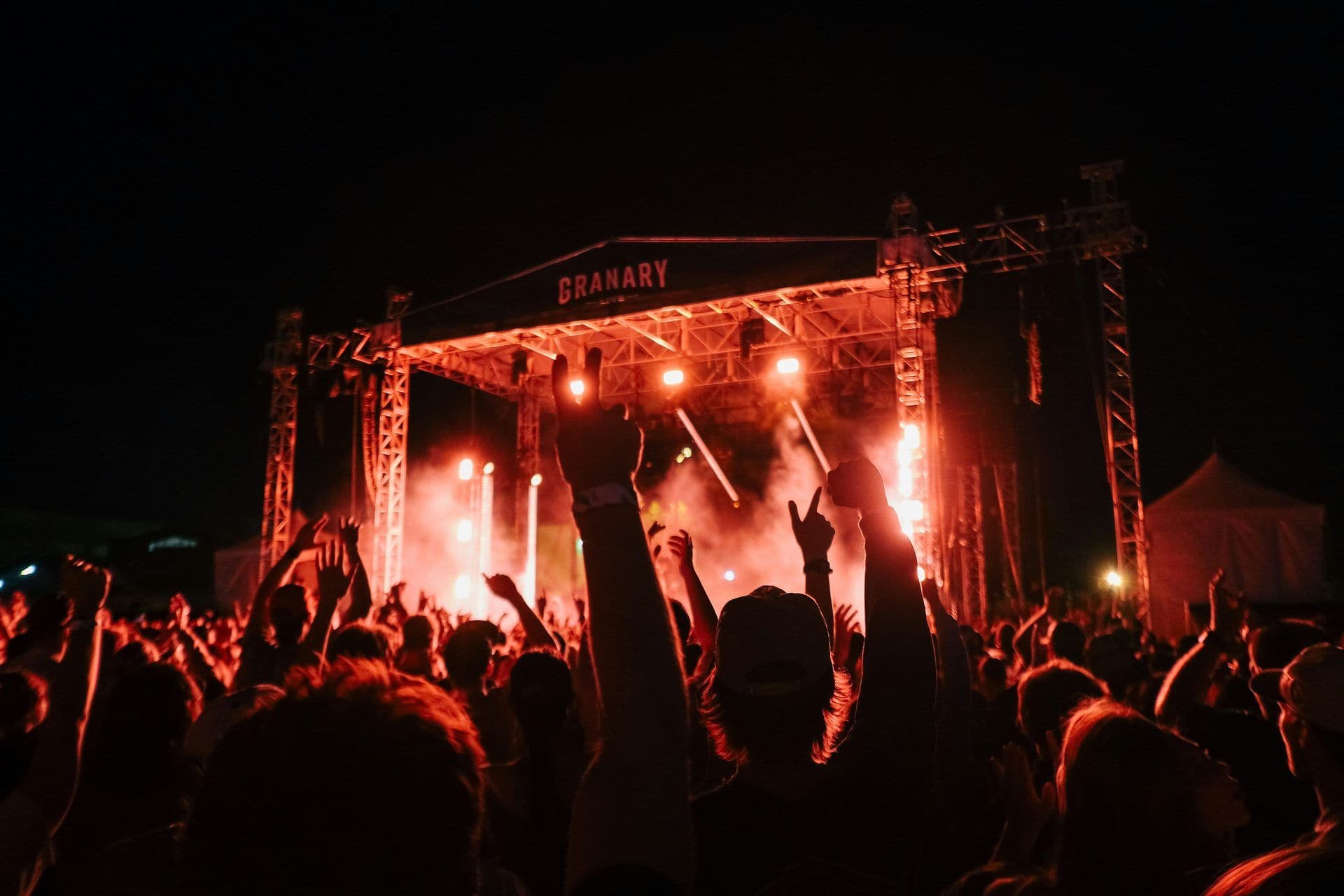 A large crowd with raised hands enjoys a night concert at an outdoor stage lit with red lights