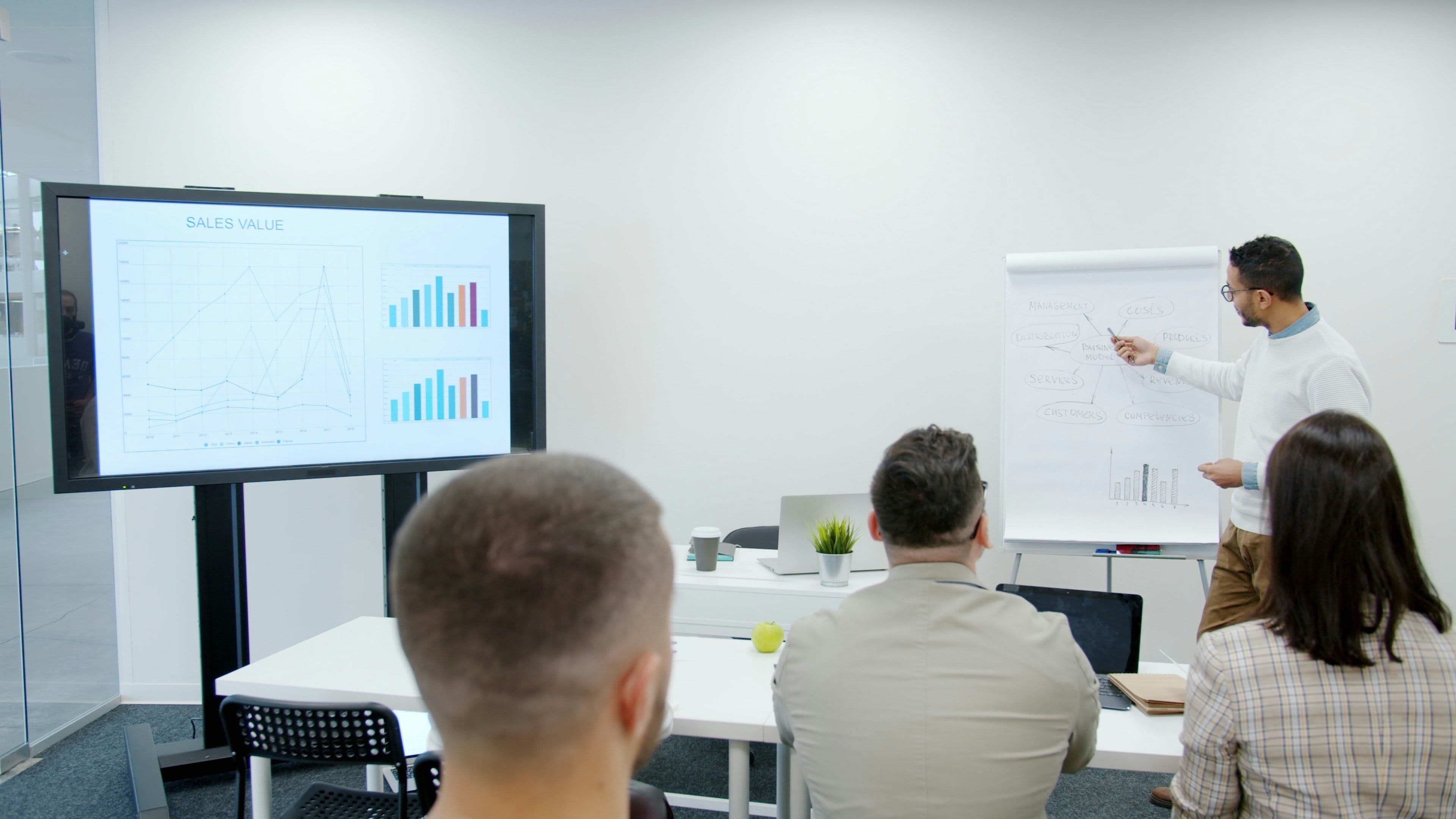 A presenter explains a business model on a flipchart during a corporate seminar in a modern meeting room.