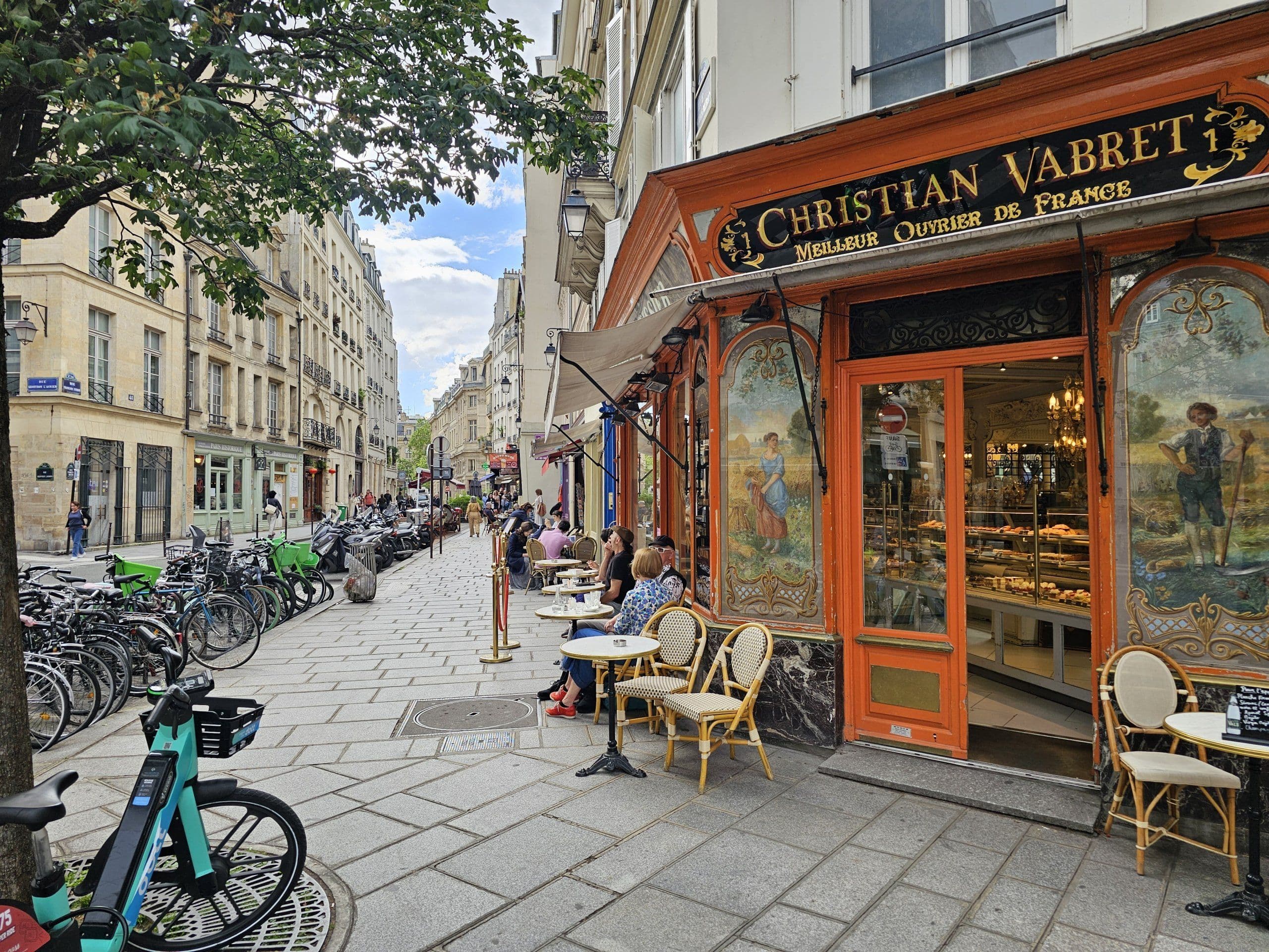A charming Parisian street with Christian Vabret patisserie, outdoor cafe seating, ideal for a business lunch.