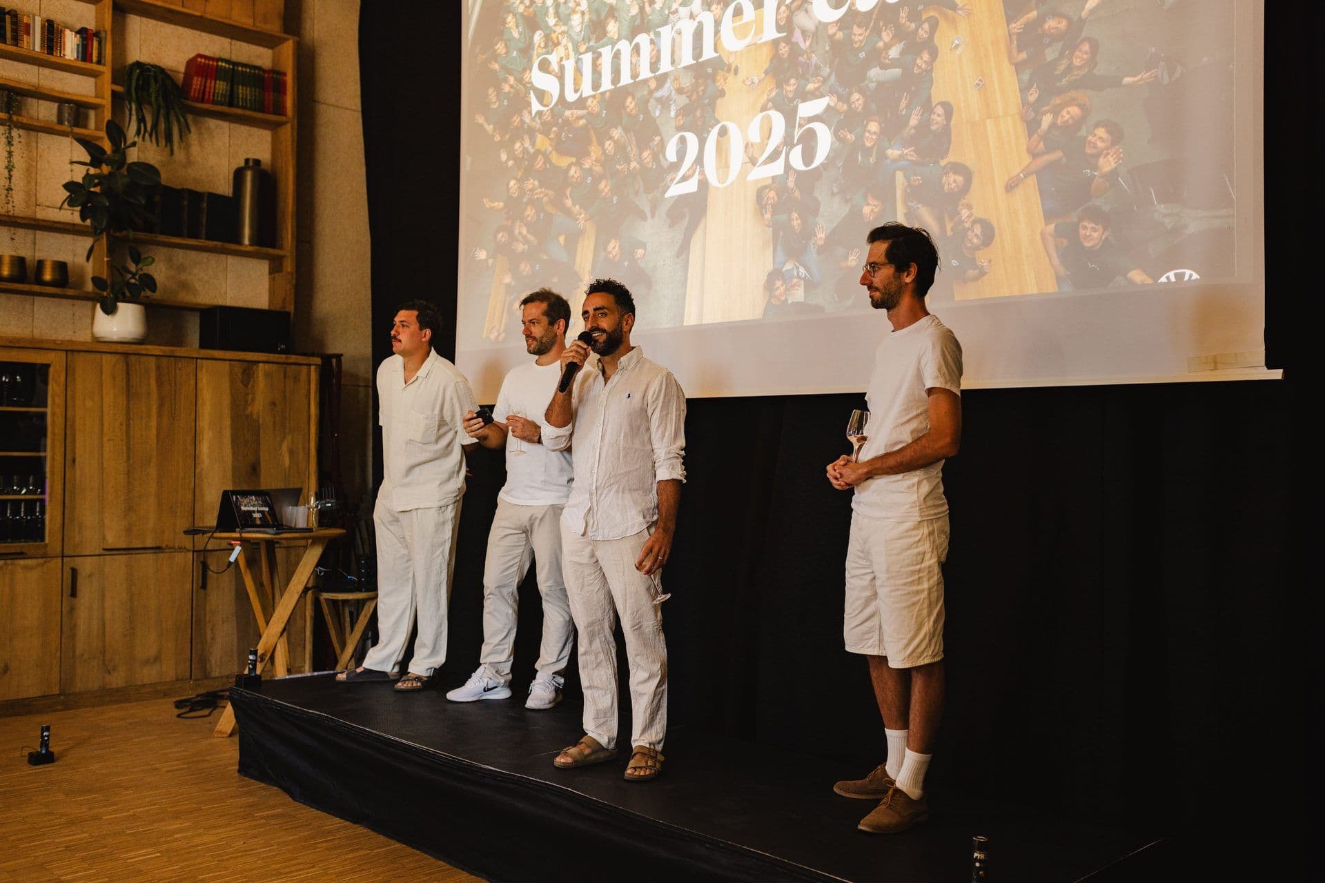 Four men on a stage present at a corporate event, with a large screen displaying "Summer Camp 2025" and a