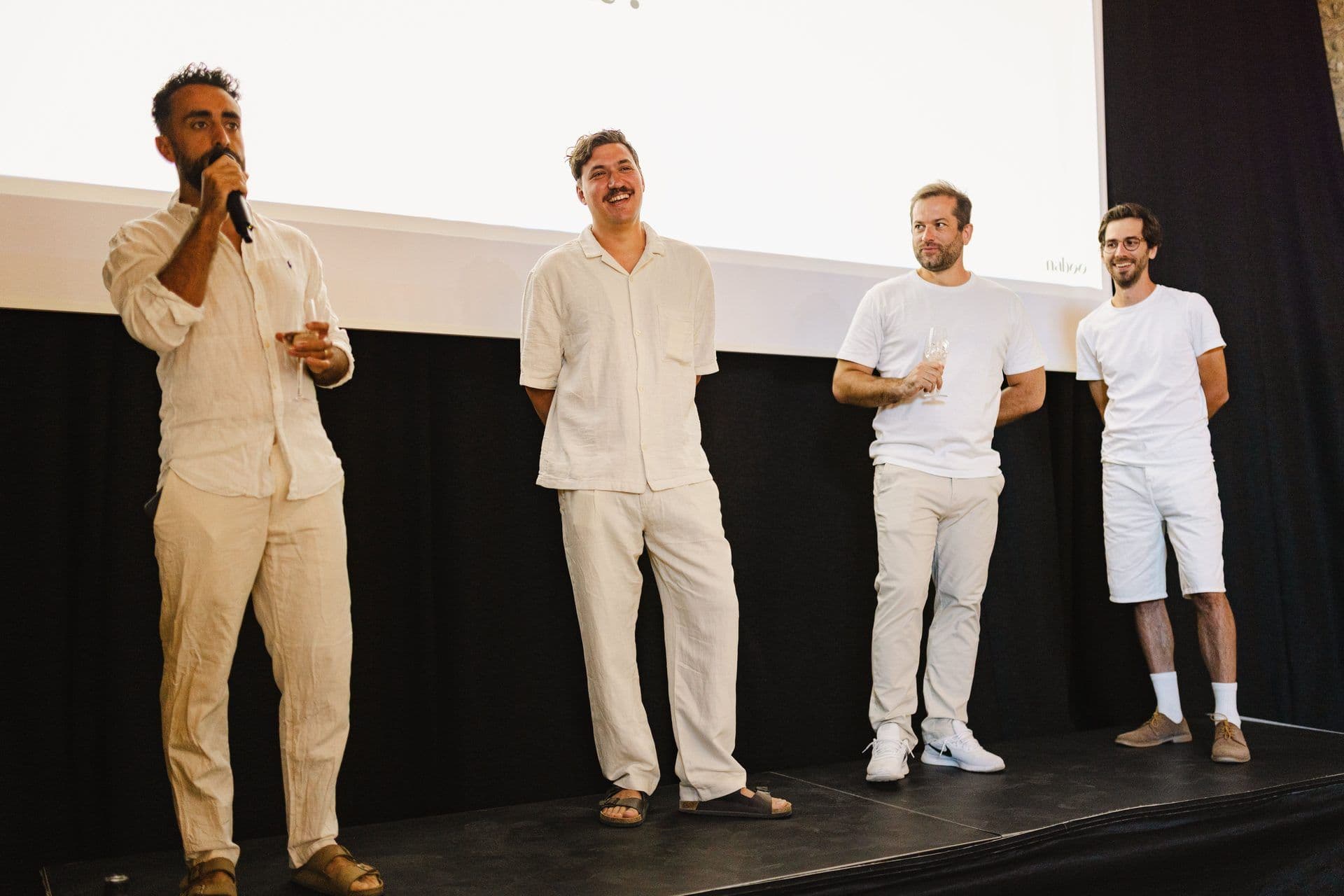 Four men on a stage, one speaking into a microphone at a corporate seminar or town hall event.