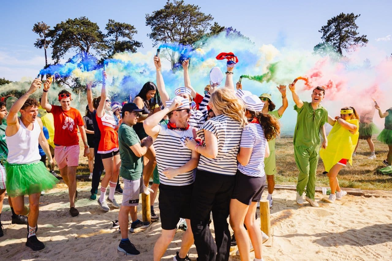 A diverse group enjoys an outdoor team building activity with colorful smoke flares and fun costumes.