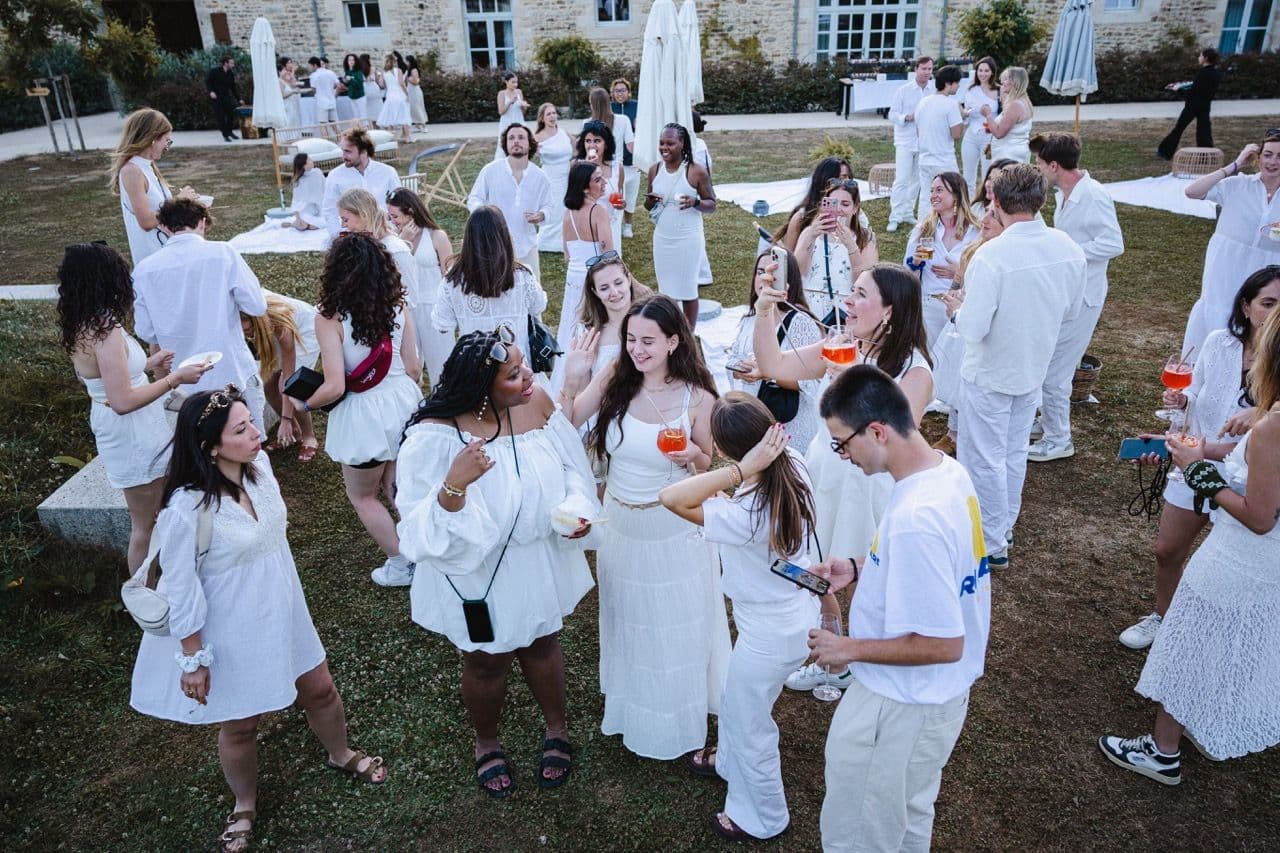 Guests in white attire mingle at an outdoor corporate cocktail reception on a lawn with a historic building.