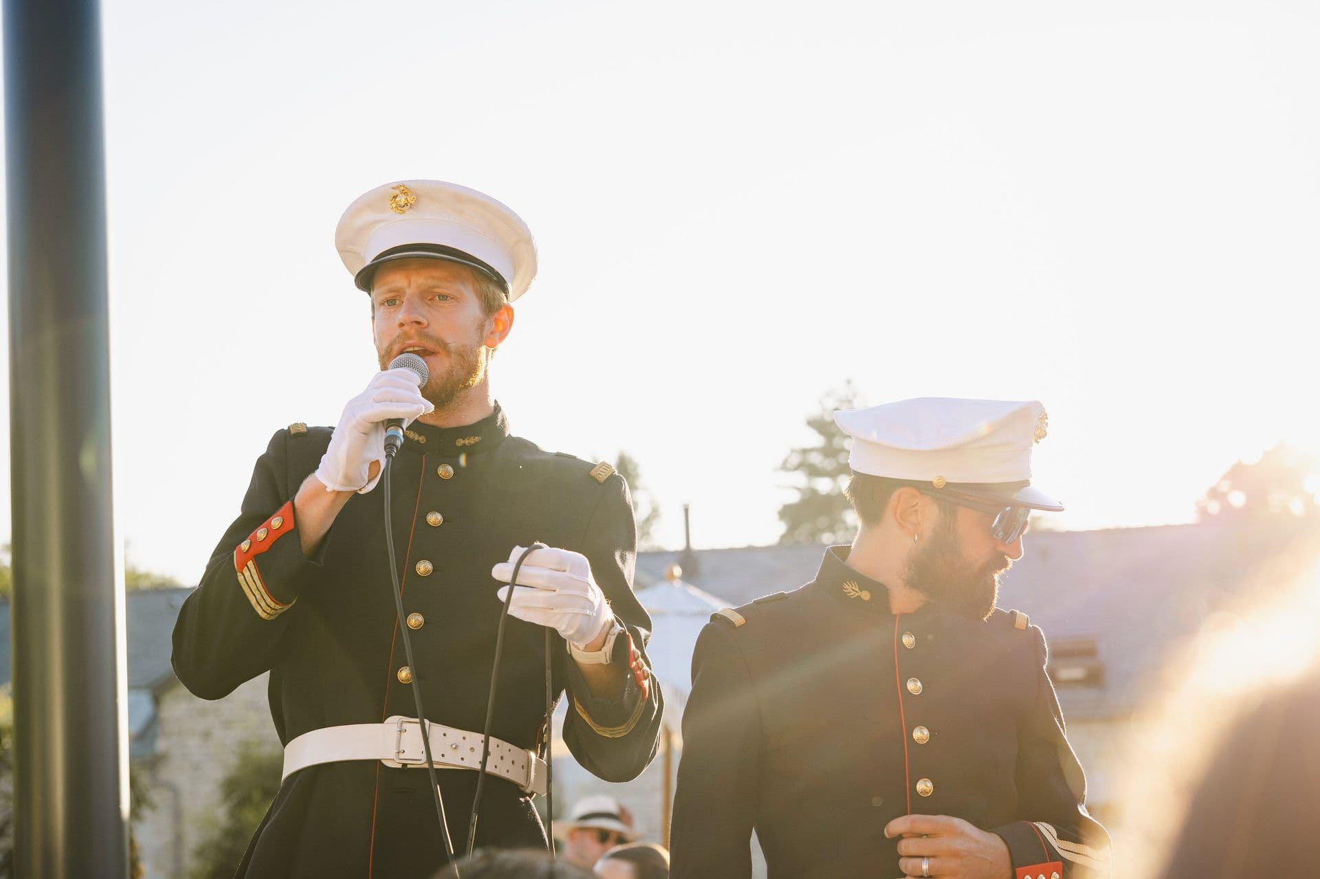 Two men in ceremonial uniforms perform outdoors with a microphone for a corporate event.