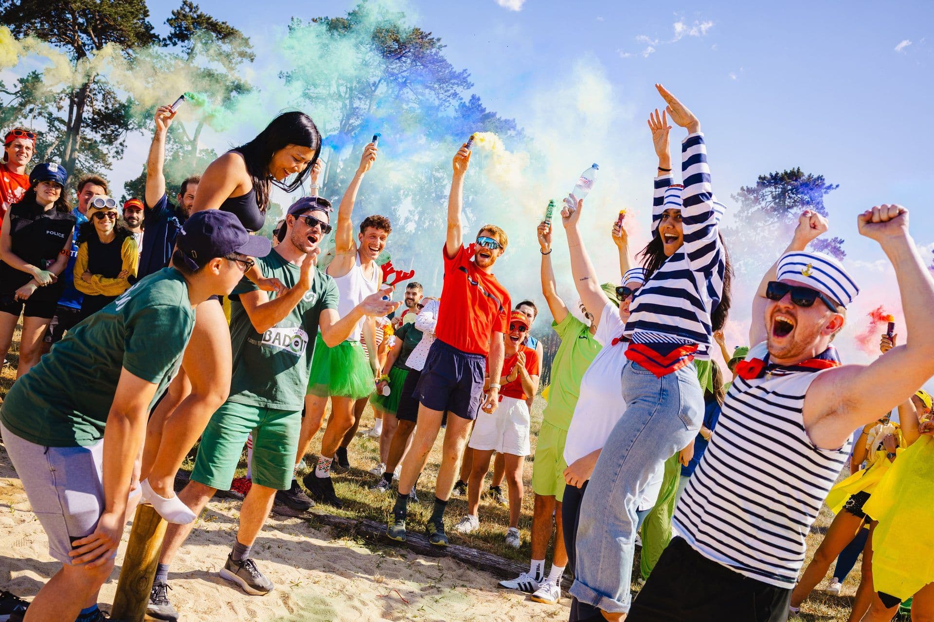 A large group of excited colleagues celebrate outdoors with colorful smoke flares during a team building