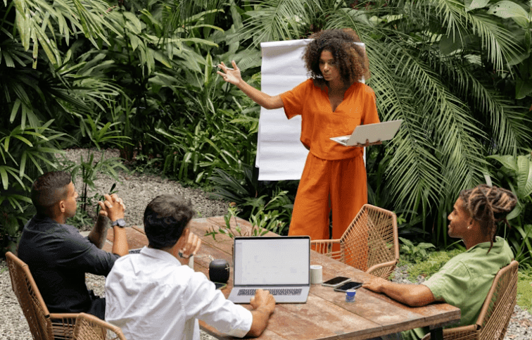 An outdoor corporate workshop with a woman presenting to a team at a wooden table amidst tropical plants.