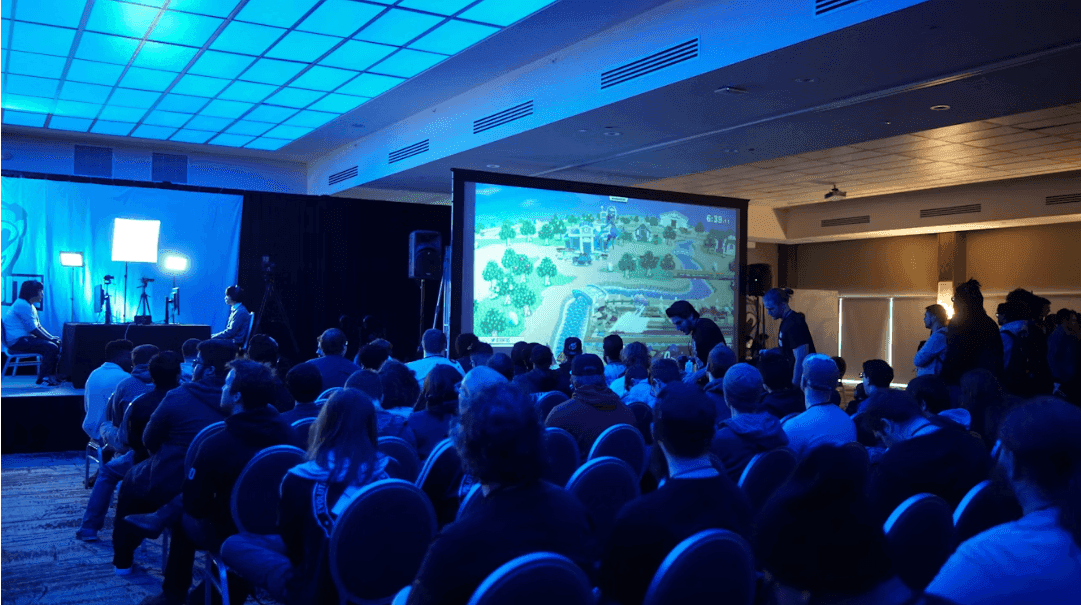 A large group attends a corporate seminar in a conference space with a stage and projection screen.