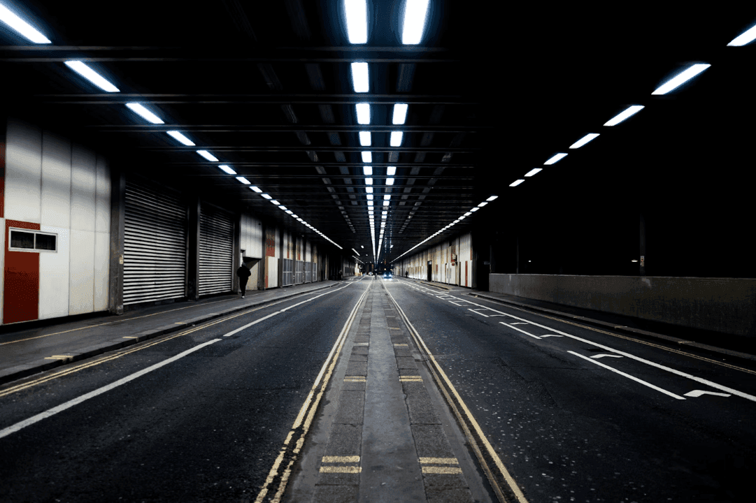 A long, dark urban tunnel with bright overhead fluorescent lights, an empty road, and a lone person walking.