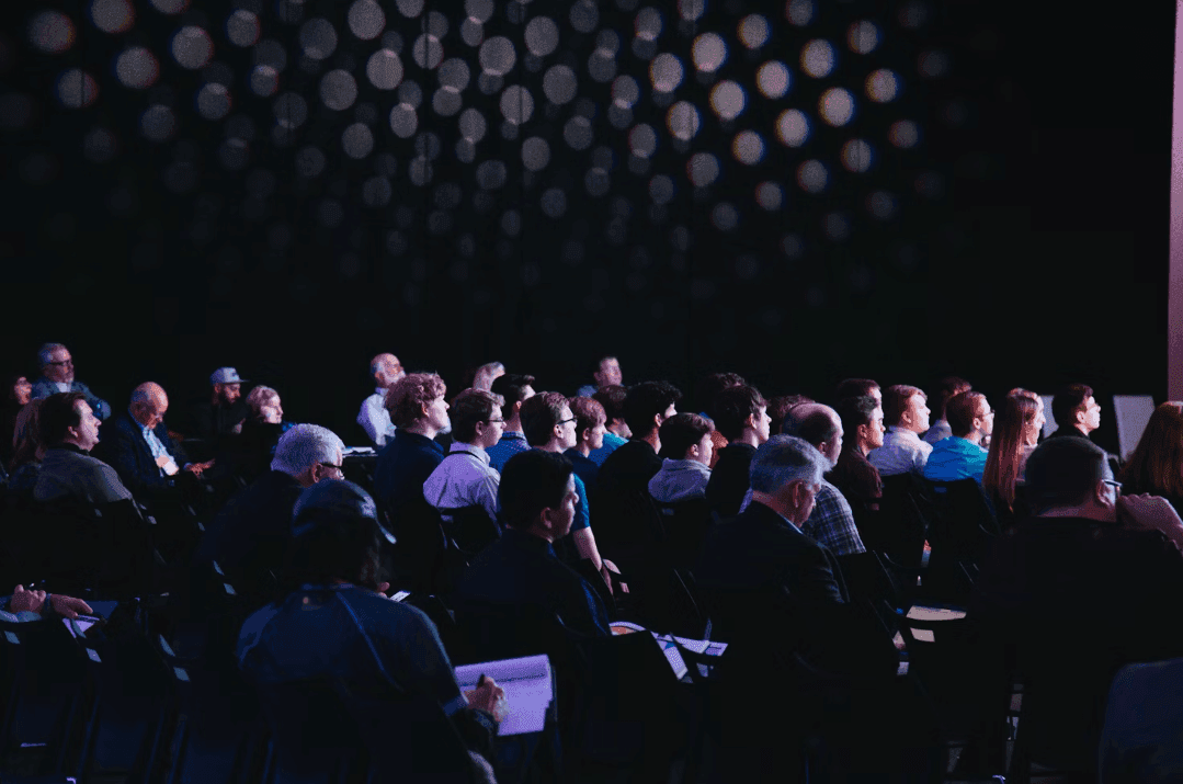 Attendees at a corporate seminar or large group workshop in a dark conference space.