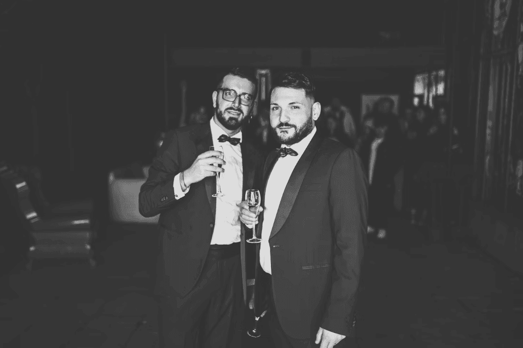 Two men in tuxedos holding champagne flutes at a sophisticated corporate dinner or cocktail reception.
