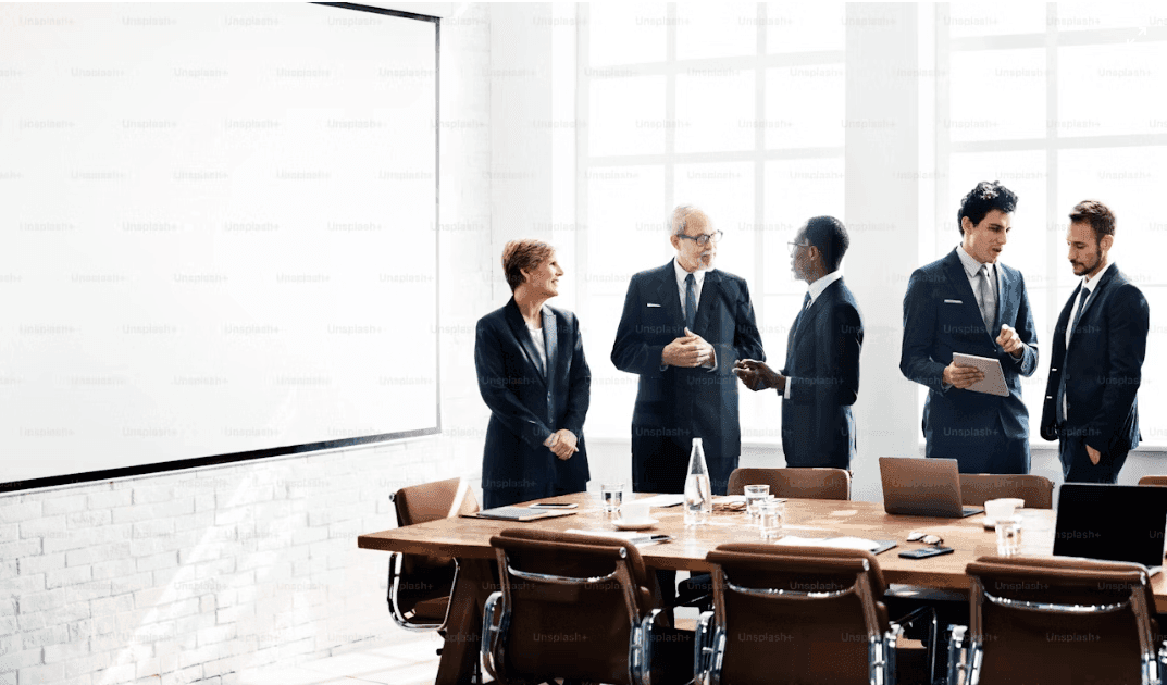 Five business professionals discussing in a modern conference room with a large table and screen.