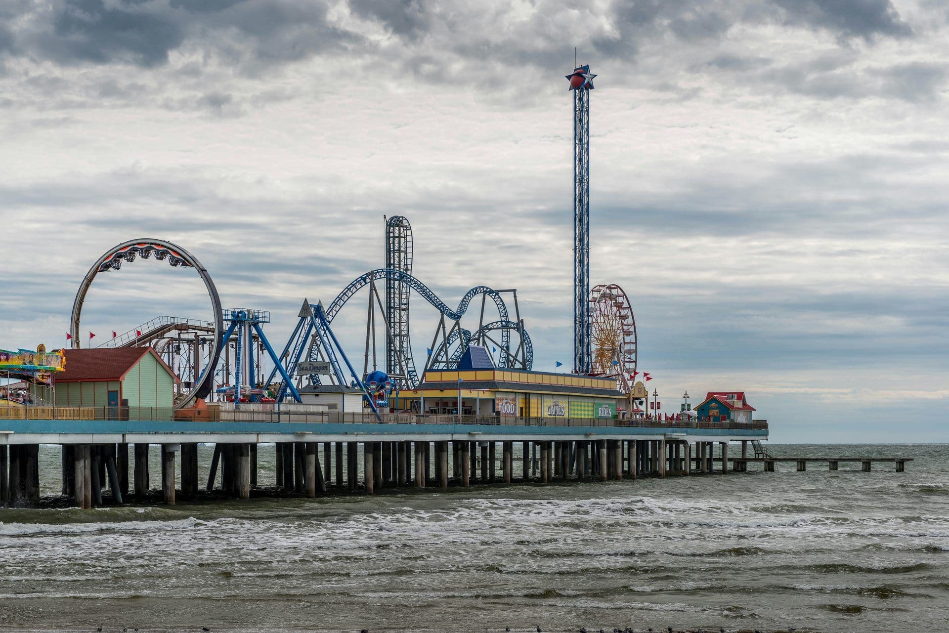 Galveston Island Historic Pleasure Pier with roller coasters and rides over the ocean under a cloudy sky.