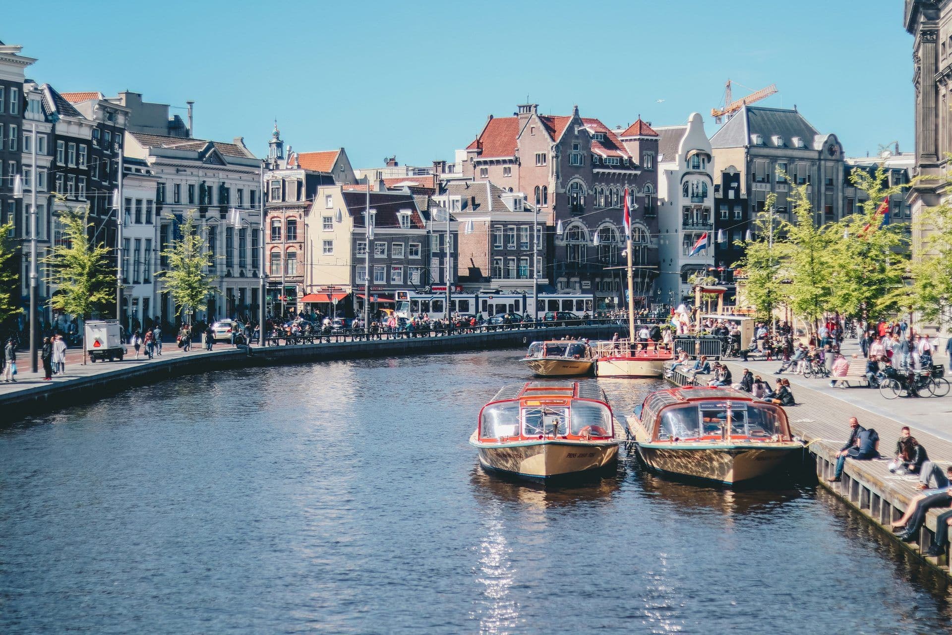 Amsterdam canal scene with traditional gabled buildings, tour boats, and people enjoying the waterfront.