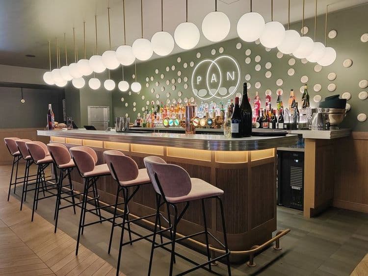 A modern bar area featuring pink velvet stools, a fully stocked bar, and spherical pendant lights.
