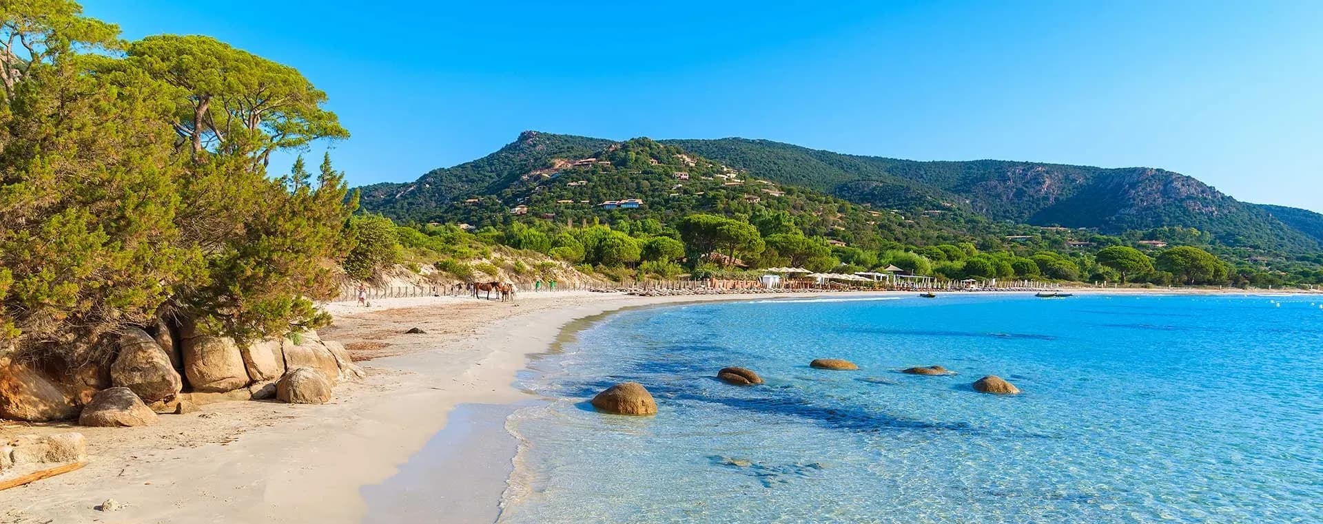 Idyllic waterfront venue in Corsica with a sandy beach, clear blue sea, and lush green mountains.