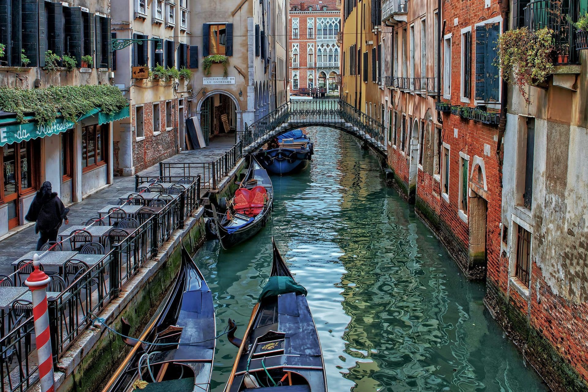 Gondolas moored on a narrow canal in Venice, Italy, with historic buildings and an outdoor restaurant.
